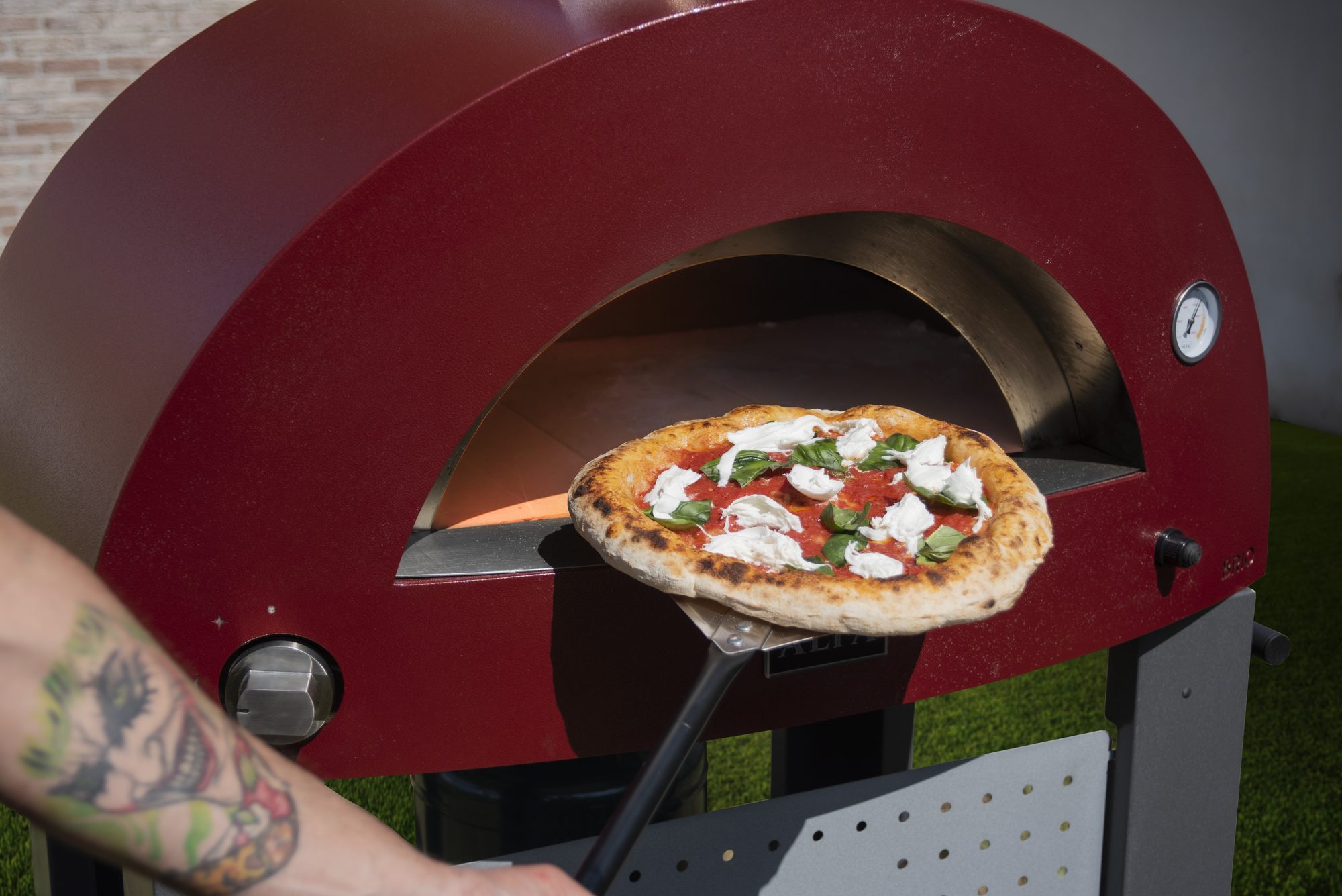 A wood-fired pizza on a peel coming out of a red Alfa oven