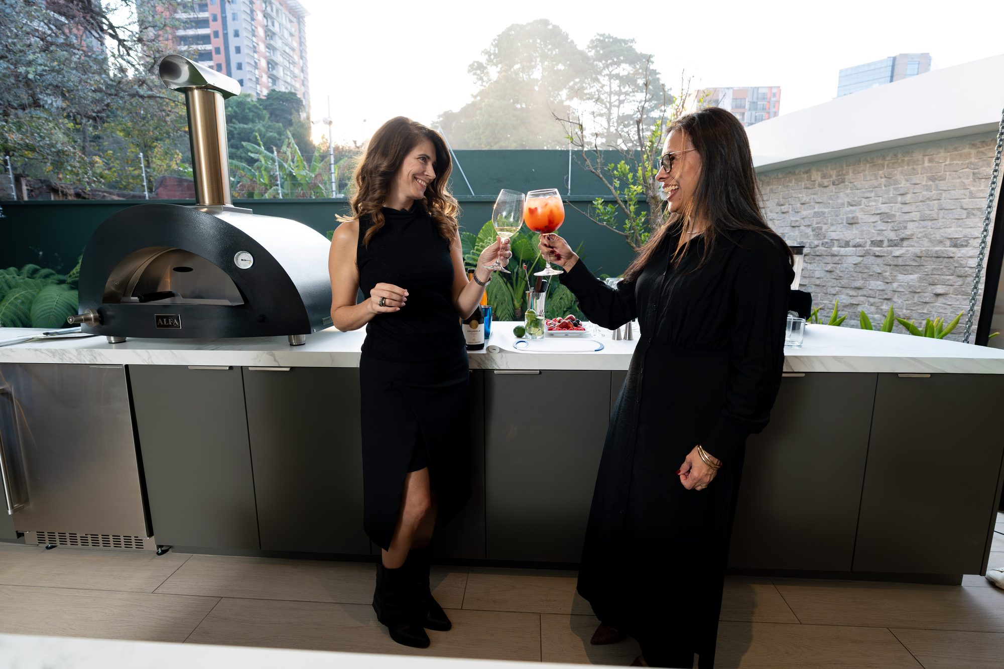 Two guests toasting with drinks beside an Alfa oven in an outdoor kitchen
